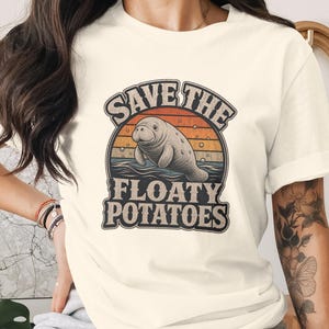 May include: A cream-coloured short-sleeve t-shirt featuring a graphic print of a manatee swimming in water. Above and below the manatee, the text reads "SAVE THE FLOATY POTATOES" in a bold, dark font, set against a retro-style orange and blue sunset.