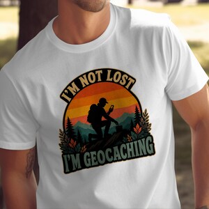 May include: A white short-sleeved t-shirt featuring a graphic of a hiker kneeling in a mountainous landscape at sunset. The text reads I'M NOT LOST I'M GEOCACHING in bold lettering. The design includes silhouettes of pine trees and foliage.