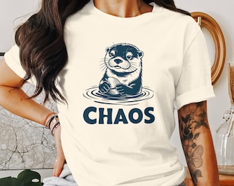 Puede incluir: Una camiseta blanca con un gráfico de una nutria sosteniendo una roca en el agua. El texto "CHAOS" está impreso debajo de la nutria.