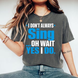 May include: A charcoal gray short-sleeve t-shirt featuring the white and bright blue text I DON'T ALWAYS Sing ... OH WAIT YES I DO. The shirt has a crew neckline and a relaxed fit, showcasing a bold graphic design printed on the front center.