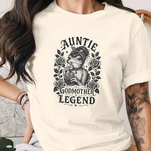 May include: A cream-coloured short-sleeve t-shirt featuring a black graphic print of a frog wearing a bandana and drinking from a glass. The text reads AUNTIE GODMOTHER LEGEND, surrounded by floral rose illustrations and decorative leaves.