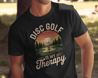 May include: Black t-shirt with a graphic design featuring a disc golf course scene under a sunset. The text "Disc Golf is my Therapy" is arched above the scene and below it. The shirt is made of a soft material.