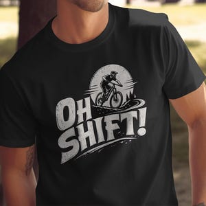 May include: A black short-sleeve t-shirt featuring a white graphic print of a mountain biker riding on a trail in front of a large circular moon. The text "OH SHIFT!" is printed in large, distressed white capital letters below the graphic.