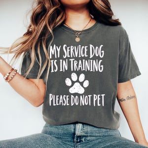 May include: A charcoal gray short-sleeve t-shirt featuring white distressed text that reads MY SERVICE DOG IS IN TRAINING PLEASE DO NOT PET, centered above and below a white graphic of a single paw print with a textured, worn-in appearance.