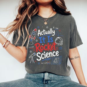 May include: Dark grey t-shirt featuring the phrase "Actually It Is Rocket Science" in red, white, and blue. The design includes a rocket, astronaut helmet, telescope, and equations. A casual, science-themed tee.