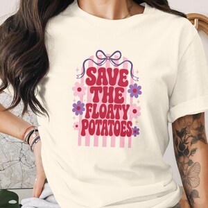 May include: A cream-coloured t-shirt featuring a graphic design with the text "SAVE THE FLOATY POTATOES" in red, stylised lettering. The design includes a purple bow at the top, pink vertical stripes, and small purple and pink flower accents.