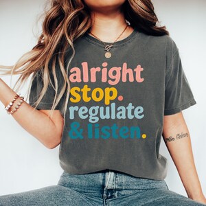 May include: A charcoal grey short-sleeve t-shirt featuring the text "alright stop. regulate & listen." printed in stacked lines. The word "alright" is in pink, "stop." is in yellow, "regulate" is in light blue, and "& listen." is in teal.