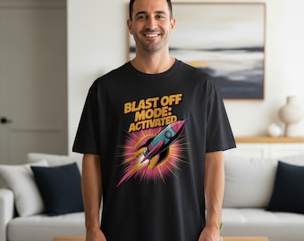 Może przedstawiać: Czarny t-shirt z napisem "BLAST OFF MODE: ACTIVATED" w kolorze żółtym i grafiką rakiety z różowymi, niebieskimi i czarnymi akcentami. Rakieta otoczona jest żółtymi i różowymi liniami.