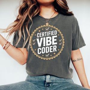 May include: Dark gray t-shirt with a circular graphic that reads "CERTIFIED VIBE CODER" in white text. The graphic has a gold border with blue stars and checkmarks. The shirt is a casual style.