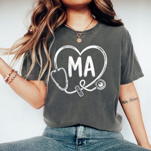 Puede incluir: Una camiseta de manga corta gris carbón con un diseño gráfico blanco de un contorno de corazón formado por un estetoscopio. Las letras MA están impresas en el centro del corazón y un pequeño icono de tirita está colocado en el tubo del estetoscopio.