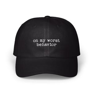 May include: A black baseball cap featuring the white embroidered text on my worst behavior centered on the front panel. The cap has a structured crown, a curved brim, and a button at the top, constructed from a solid black fabric material.