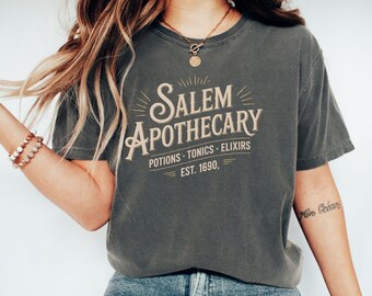May include: Dark grey t-shirt with the text "Salem Apothecary" in a vintage style, with the words "Potions, Tonics, Elixirs" and "Est. 1690" below. The shirt is a comfortable, casual style.