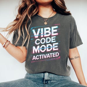 May include: Dark grey t-shirt with the text "VIBE CODE MODE ACTIVATED" in a pixelated, glitch-style design. The text is in white, blue, and pink. The shirt is a casual, comfortable style.