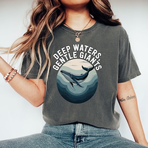 以下が含まれることがあります： 円形デザインの中に青いクジラのイラストが描かれた、濃いグレーのTシャツ。「DEEP WATERS GENTLE GIANTS」の文字は白。クジラは青い水の中を泳いでいます。