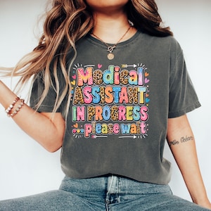 May include: A charcoal gray short-sleeve t-shirt featuring the colorful text Medical ASSISTANT IN PROGRESS... please wait. The lettering incorporates tie-dye patterns, leopard print, and solid colors, accented by small hearts and arrows.