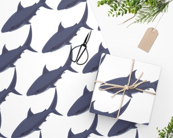 Puede incluir: Papel de regalo con un patrón repetitivo de tiburones azul oscuro sobre fondo blanco. Una caja de regalo cuadrada envuelta en el mismo papel de tiburones está atada con cuerda beige. Unas tijeras de metal negro descansan sobre el papel.