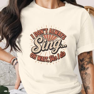 Könnte beinhalten: Ein cremefarbenes Kurzarm-T-Shirt mit einem Grafikdesign, das ein Sonnenstrahlen-Motiv und den Text "I DON'T ALWAYS Sing... OH WAIT, Yes I do" in einer Kombination aus kräftigen Serifenschriften und Schreibschrift in Orange und Schwarz zeigt.