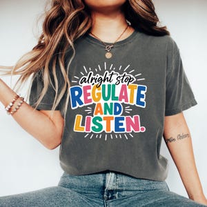 Op de afbeelding: Een antracietgrijs T-shirt met korte mouwen en een kleurrijke grafische print met de tekst "alright stop REGULATE AND LISTEN." De letters gebruiken een mix van blauw, groen, oranje, geel en roze met witte omlijningen en uitstralende lijnen.