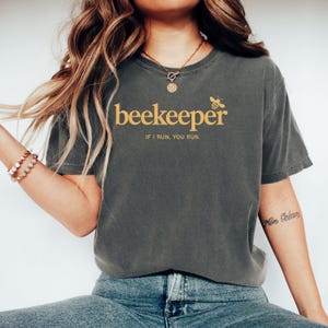 May include: Dark gray t-shirt with the word "beekeeper" in gold lettering, along with a bee graphic and the phrase "IF I RUN, YOU RUN." The shirt has short sleeves and a crew neck.