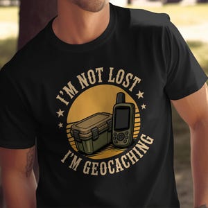 May include: A black t-shirt featuring a graphic print of a tan storage container and a handheld GPS device centred over a yellow sun icon. The text "I'M NOT LOST" appears in an arch above the graphic, and "I'M GEOCACHING" is printed below in a bold font.