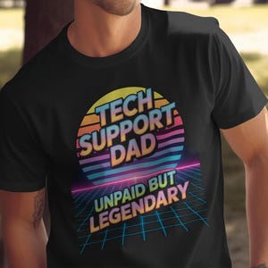 Può includere: T-shirt nera con un design retrò. La grafica presenta le parole "TECH SUPPORT DAD" in un design circolare colorato, con "UNPAID BUT LEGENDARY" sotto. Il design ha un'estetica anni '80.