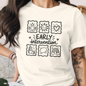 May include: A cream-colored t-shirt featuring a black graphic print with the text "EARLY intervention" centered among six square icons depicting hands holding a star, a sun, a heart, building blocks, a speech bubble, and a rainbow.