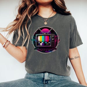 May include: A charcoal grey short-sleeve t-shirt featuring a circular graphic of a vintage television set with a colourful test pattern screen, antennas, and a digital glitch effect set against a vibrant pink and purple sunset background.