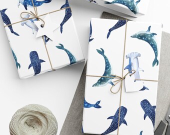 Puede incluir: Papel de regalo blanco con un patrón repetido de ballenas, delfines y tiburones martillo azules y blancos. El papel está atado con cuerda y tiene una etiqueta de regalo blanca.