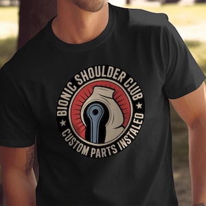 May include: Black t-shirt featuring a circular graphic with the text "BIONIC SHOULDER CLUB CUSTOM PARTS INSTALLED." The graphic depicts a stylized shoulder joint in white, red, and blue. The shirt is short-sleeved.