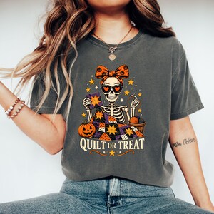 May include: A charcoal grey t-shirt featuring a graphic of a skeleton wearing a pumpkin-patterned bow and heart-shaped sunglasses. The skeleton is quilting a colourful star-patterned quilt with a pumpkin and yarn nearby. Text reads QUILT OR TREAT.