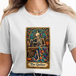 Könnte beinhalten: Ein weißes Kurzarm-T-Shirt mit einem rechteckigen Grafikdruck eines Skeletts an einer Nähmaschine. Das Design ähnelt einer Tarotkarte mit dem Text The Quilter am unteren Rand, einem bunten Patchwork-Quilt, Kerzen und einem Kürbis als Motiv.
