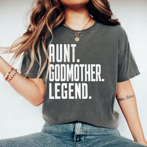 Könnte beinhalten: Ein anthrazitfarbenes T-Shirt mit kurzen Ärmeln und Rundhalsausschnitt, das auf der Brust den weißen, im Used-Look gedruckten Text AUNT. GODMOTHER. LEGEND. in einer fetten, serifenlosen Schrift zeigt. Das Kleidungsstück hat eine lockere Passform.