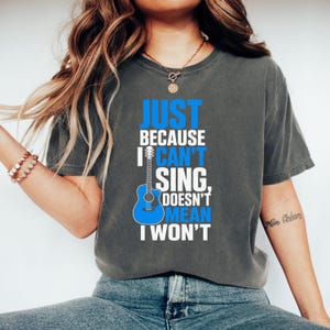 Könnte beinhalten: Ein anthrazitfarbenes Kurzarm-T-Shirt mit einer blauen Akustikgitarre als Grafik und weißem sowie blauem Text, der lautet: JUST BECAUSE I CAN'T SING, DOESN'T MEAN I WON'T. Das T-Shirt hat einen Rundhalsausschnitt und eine lockere Passform aus Baumwolle.