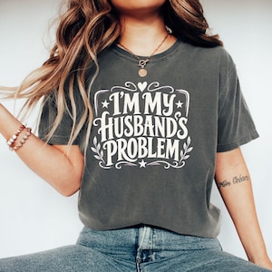 Könnte beinhalten: Ein anthrazitfarbenes Kurzarm-T-Shirt mit dem weißen Schriftzug I'M MY HUSBAND'S PROBLEM in einer dekorativen Schriftart, akzentuiert durch kleine Sterne, ein Herz und Blattmotive. Das T-Shirt hat einen Rundhalsausschnitt und eine lockere Passform.