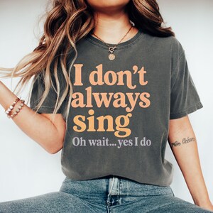 May include: A charcoal grey short-sleeve t-shirt featuring the orange text "I don't always sing" and the smaller grey text "Oh wait... yes I do" printed on the front. The shirt has a crew neckline and a relaxed fit with a vintage-washed appearance.