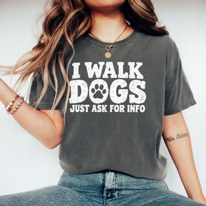 Könnte beinhalten: Ein anthrazitfarbenes Kurzarm-T-Shirt mit weißem, abgenutztem Text, der I WALK DOGS JUST ASK FOR INFO lautet, wobei ein Pfotenabdruck den Buchstaben O im Wort DOGS ersetzt. Das T-Shirt hat einen Rundhalsausschnitt und eine lockere Passform.