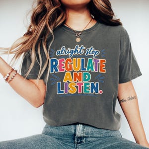 Op de afbeelding: Een antracietgrijs t-shirt met korte mouwen en de meerkleurige tekst "alright stop REGULATE AND LISTEN." De afbeelding gebruikt een verweerd, getextureerd lettertype in tinten blauw, roze, geel, oranje en groenblauw, gecentreerd op de borst.