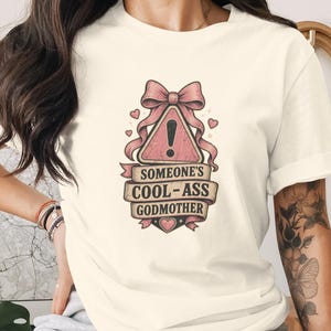 May include: A cream-coloured t-shirt featuring a graphic design of a pink bow above a triangular warning sign with an exclamation point. Below this, a banner contains the text SOMEONE'S COOL-ASS GODMOTHER, with a small heart at the bottom.