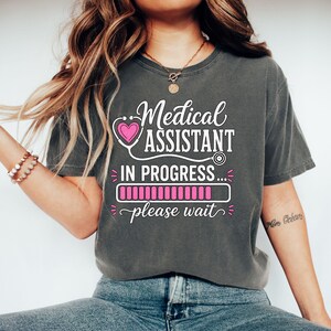 Puede incluir: Una camiseta de manga corta gris carbón con texto en blanco y rosa que dice Medical ASSISTANT IN PROGRESS... please wait. El diseño incluye un gráfico de estetoscopio con un corazón rosa y una barra de carga rosa.