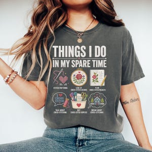 May include: Charcoal grey t-shirt with the words "THINGS I DO IN MY SPARE TIME" and cross-stitch themed illustrations. The design includes images of stitch patterns, cross-stitch designs, projects, and supplies. The phrase "I love cross stitch" is also included.
