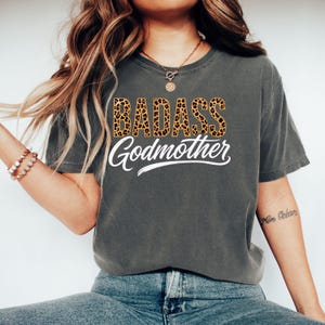 Op de afbeelding: Een antracietgrijs T-shirt met korte mouwen met de tekst BADASS in een bruin en zwart luipaardpatroon, gevolgd door het woord Godmother in een wit cursief lettertype, gecentreerd op de voorkant van het kledingstuk.