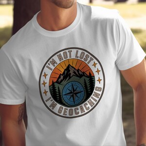 Puede incluir: Una camiseta blanca de manga corta con un gráfico circular en el pecho. El diseño muestra una cordillera, pinos y una rosa de los vientos bajo un cielo al atardecer, rodeado por el texto I'M NOT LOST I'M GEOCACHING y pequeñas estrellas.