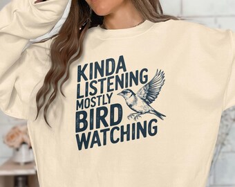 Puede incluir: Una sudadera de color crema con el texto "Kinda Listening Mostly Bird Watching" y una pequeña ilustración de un pájaro.
