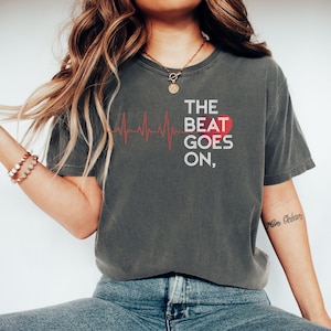 Pode incluir: T-shirt cinza escuro com o texto "THE BEAT GOES ON," uma linha vermelha de batimento cardíaco e um coração vermelho. A camisa é de estilo casual.