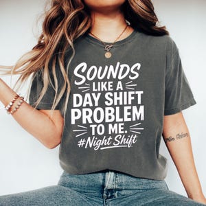 Puede incluir: Una camiseta de manga corta en color gris carbón con texto en blanco que dice: Sounds like a day shift problem to me. #Night Shift. La prenda tiene cuello redondo y un corte holgado, con el gráfico centrado en el pecho en una fuente negrita.