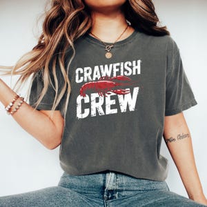 Peut inclure: Un t-shirt gris anthracite à manches courtes avec une impression graphique d'une écrevisse rouge centrée entre le texte blanc vieilli qui indique CRAWFISH CREW. Le t-shirt a une encolure ronde et une coupe décontractée au style décontracté.