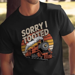 May include: Black t-shirt with a retro train graphic and the words "SORRY I TOOTED". The train is red and black, with a sunset-style backdrop. A humorous design, suitable for train enthusiasts.