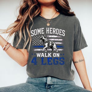 May include: A charcoal gray short-sleeve t-shirt featuring a graphic of a German Shepherd dog wearing a harness. The design includes a distressed American flag with a thin blue line and the text: SOME HEROES WALK ON 4 LEGS in white and blue.
