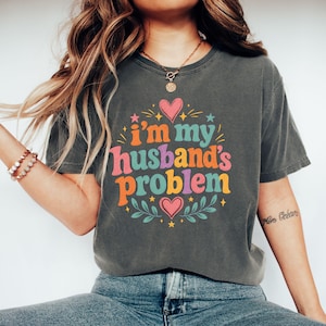 Könnte beinhalten: Ein anthrazitfarbenes Kurzarm-T-Shirt mit dem Text "i'm my husband's problem" in bunter Retro-Typografie. Das Design ist mit rosa Herzen, kleinen Sternen und grünen Blattzweigen verziert, die in einem kreisförmigen Muster angeordnet sind.