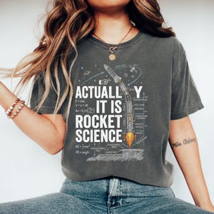 May include: Dark grey t-shirt featuring the text "ACTUALLY IT IS ROCKET SCIENCE" and a rocket diagram. The design includes equations and labels related to rocket science, with a rocket launching from a launch pad.